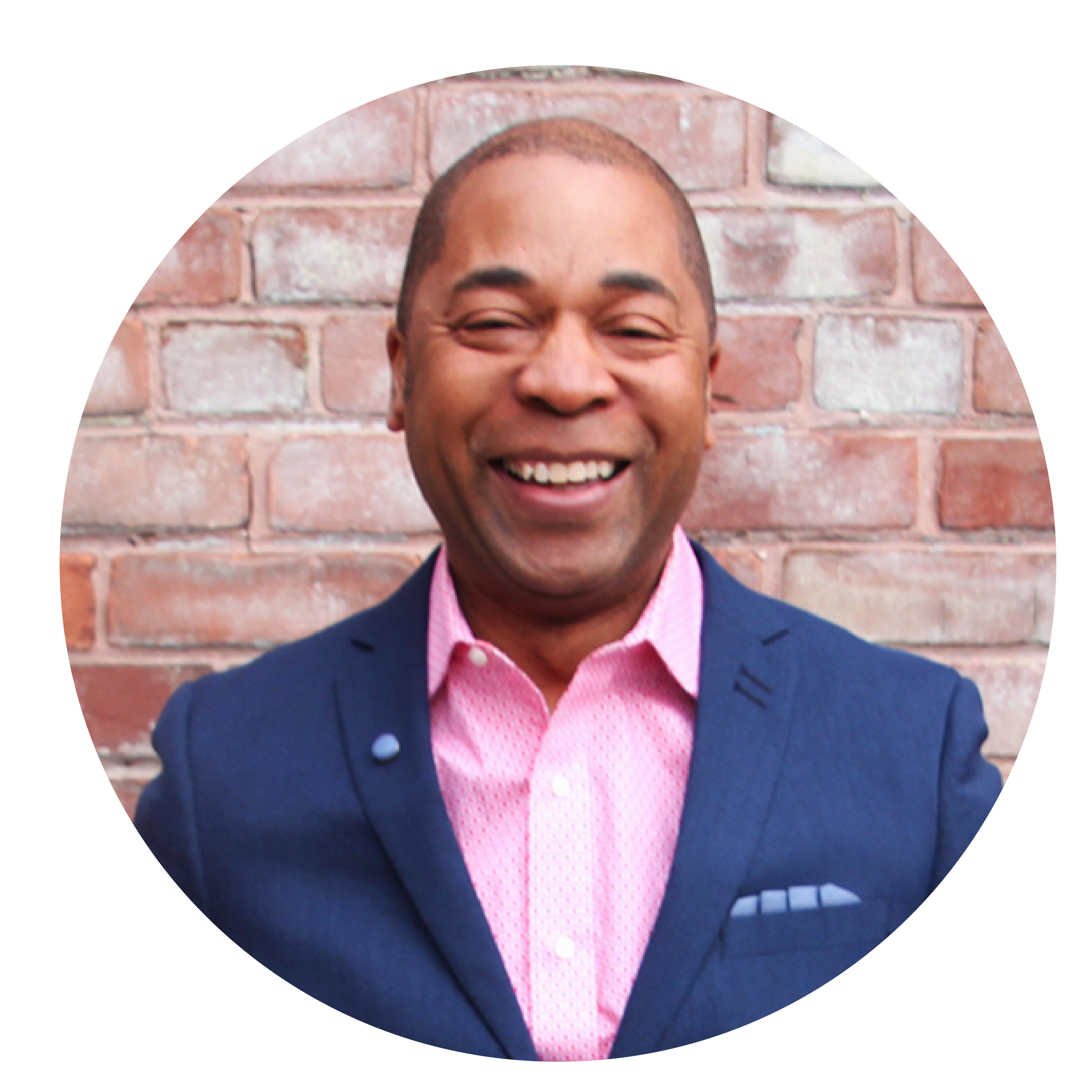 A man wearing a pink shirt and blue suit jacket smiles in front of brick wall