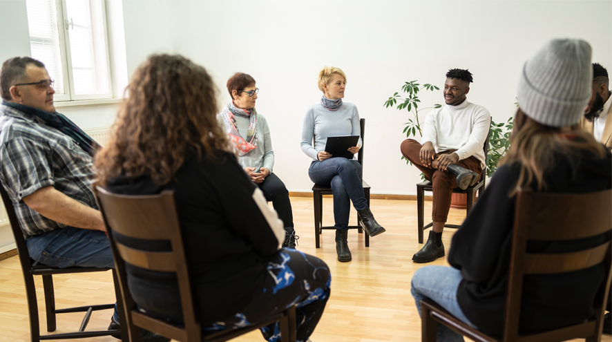 Seven adults sit on chairs in a circle, facing each other and talking