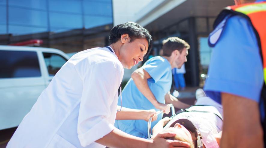 Woman with short hair and white coat helps push a gurney with patient towards hospital building