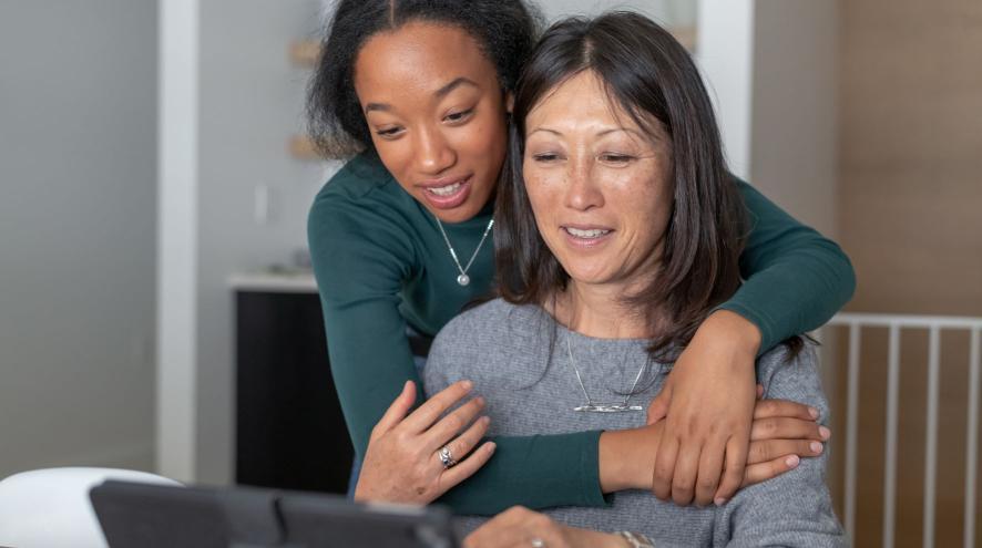 Teenager embracing midlife woman as they look at content on a tablet.