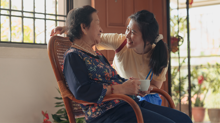 An older woman sits in a rocking chair while a teen girl speaks with her