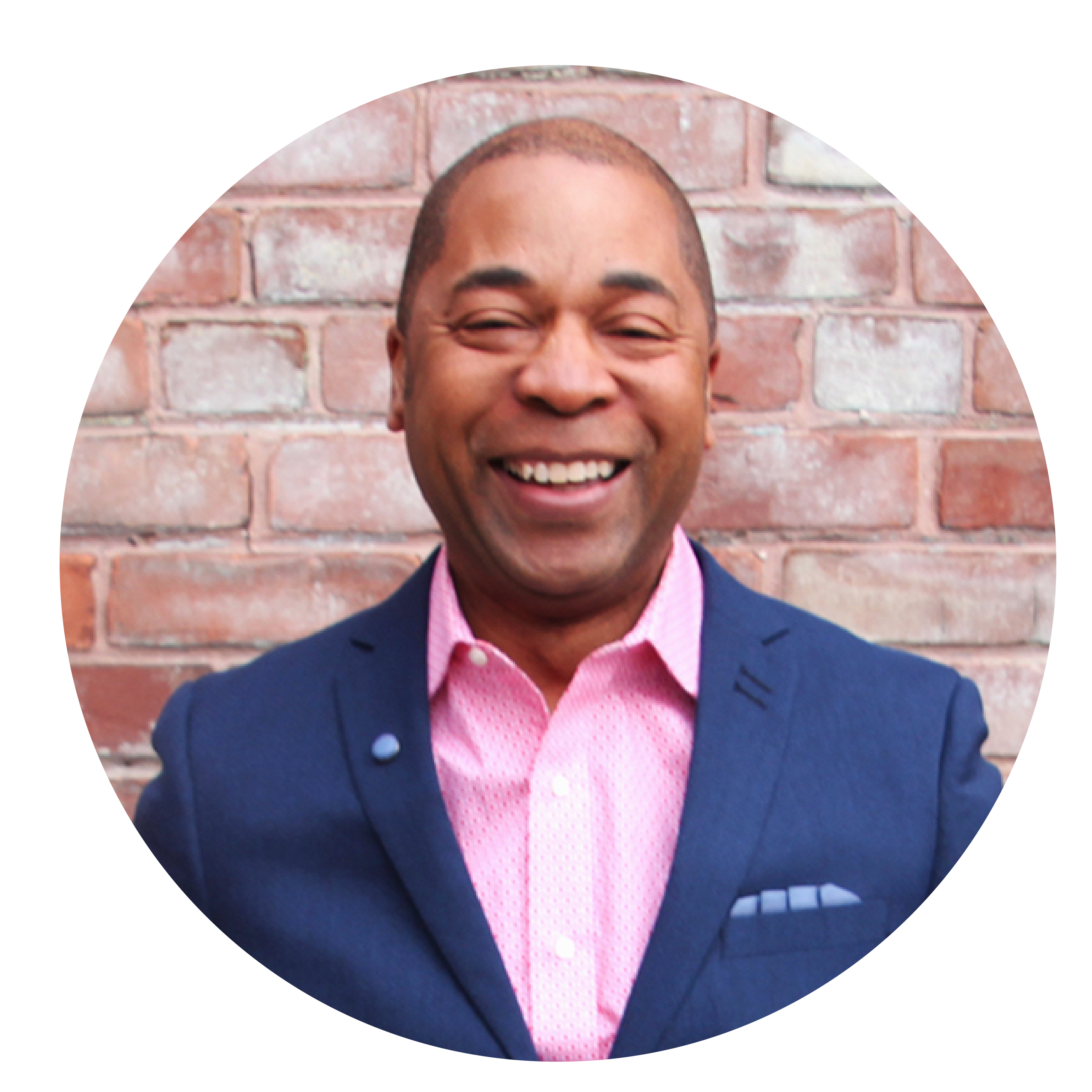 A man wearing a pink shirt and blue suit jacket smiles in front of brick wall