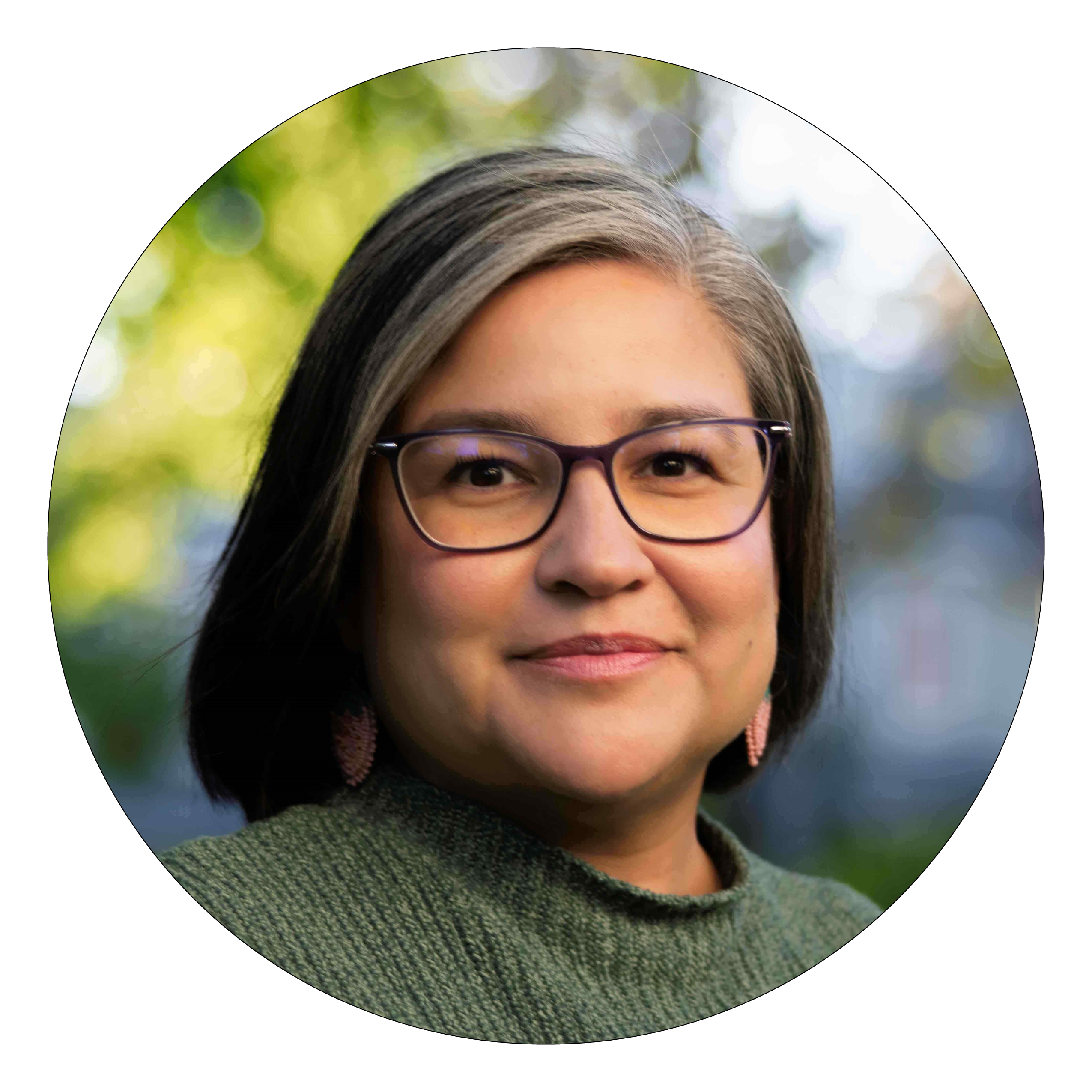 A woman with black and grey hair, green sweater and black frame glasses smiles