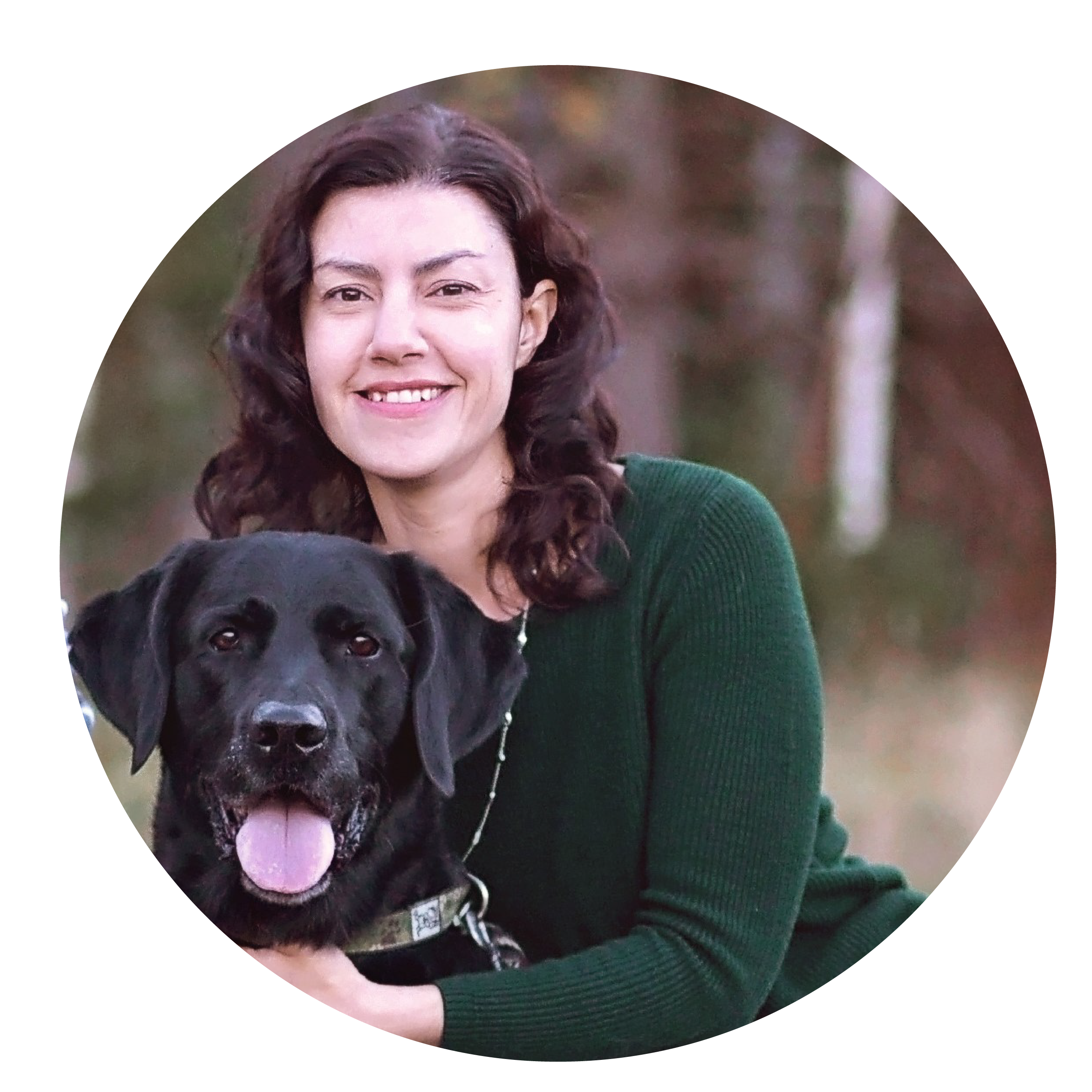 A woman with long brown hair and green sweater kneels next to a black lab dog