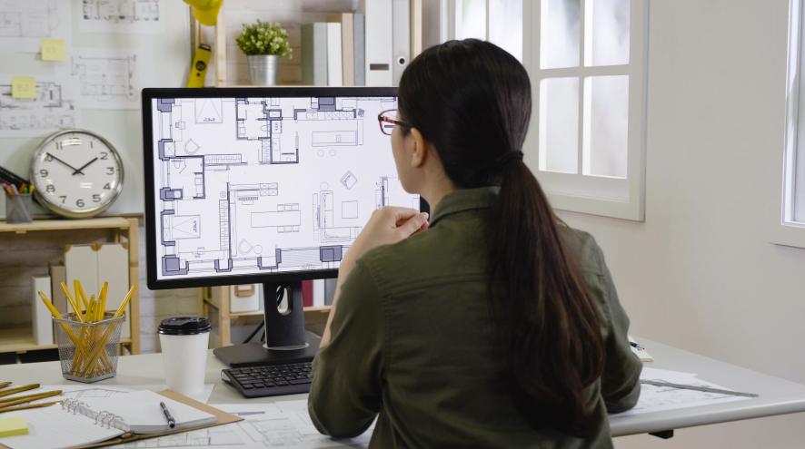 A woman with long brown hair and glasses faces a computer screen that shows a home design