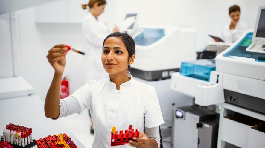 A woman in a lab coat holds up a sample tube of blood and tube rack