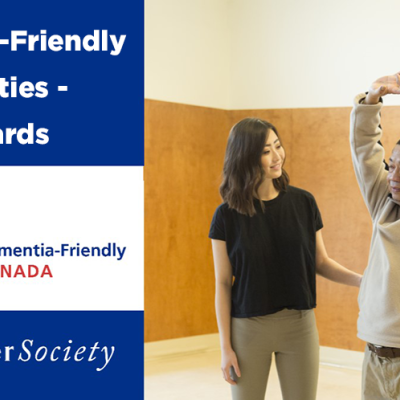 Mario, who lives with dementia, practices a stretching exercise while a friendly instructor helps. The text "Dementia-Friendly Communities - 2023 Awards" can be read to the left of Mario and the instructor.