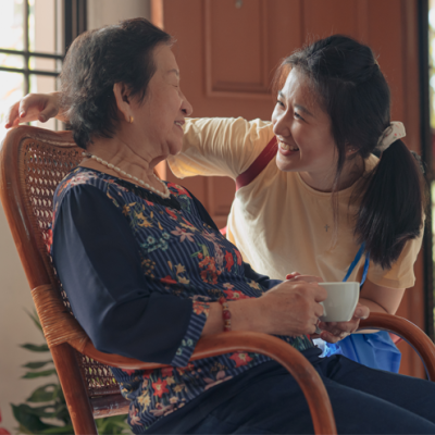 An older woman sits in a rocking chair while a teen girl speaks with her